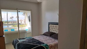 Een slaapkamer met een bed, uitzicht op een zwembad en palmbomen, en een schuifdeur naar een balkon.