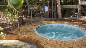 Un jacuzzi rodeado por un patio de ladrillo, barandilla metálica y un área cercada con árboles.