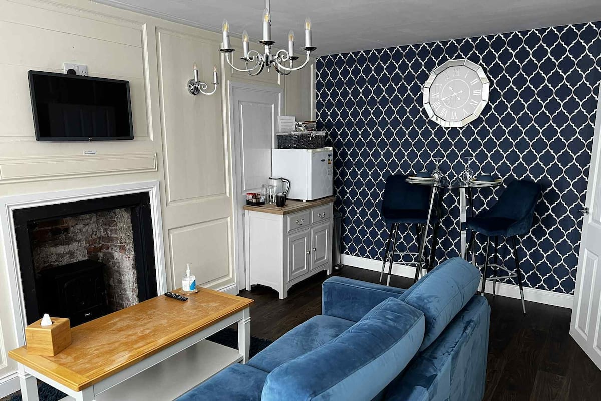 A modern living room with a fireplace, a blue sofa, a wooden coffee table, a TV mounted on the wall, and a patterned wallpaper.