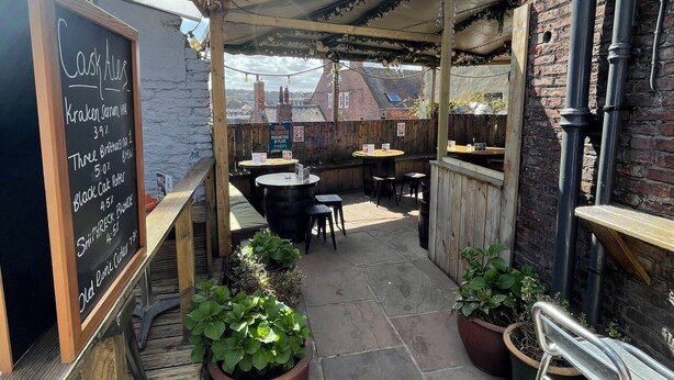 An outdoor patio area with wooden tables and chairs, a chalkboard menu, and potted plants. - The Little Angel (Whitby)