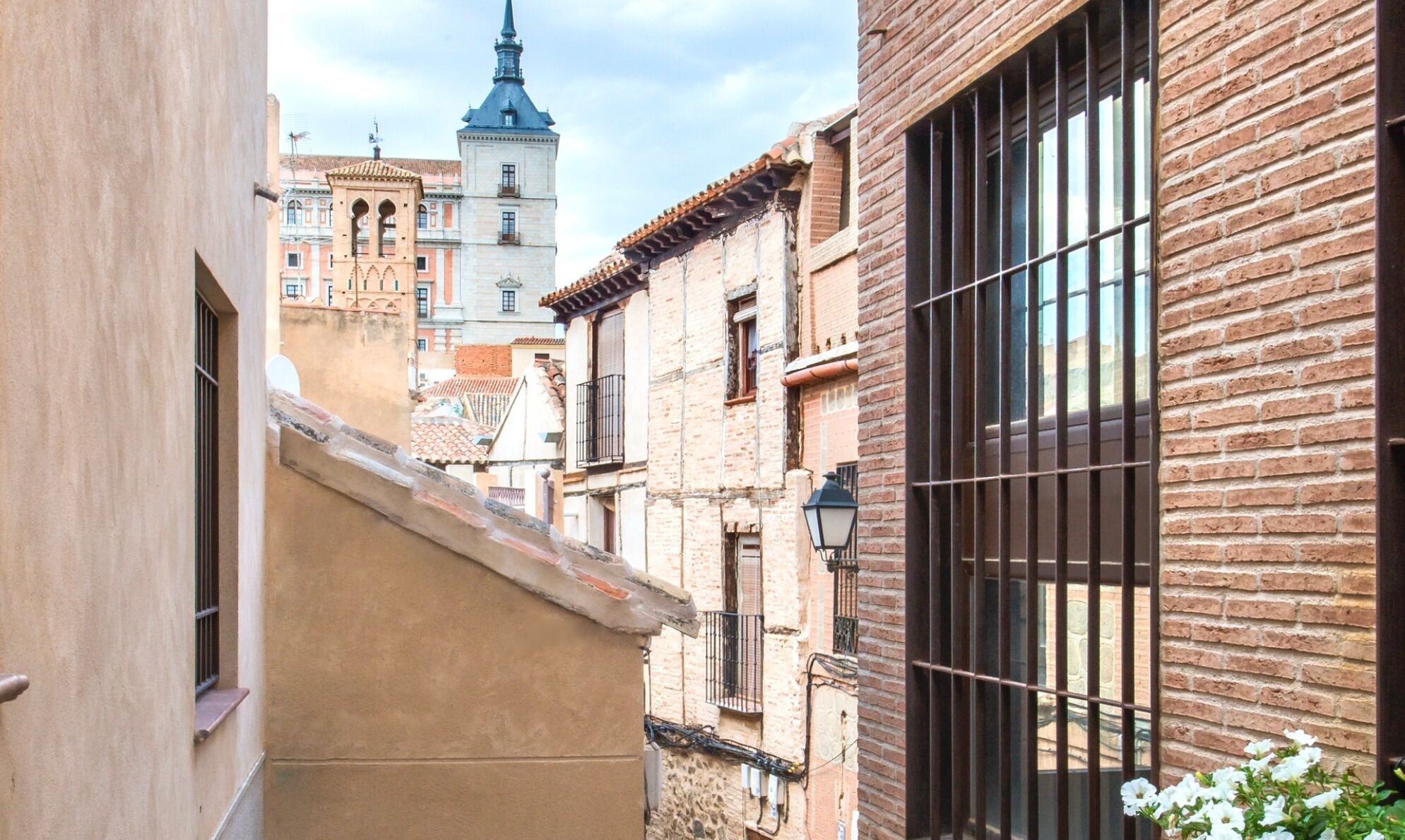 A narrow alleyway with buildings on either side, one with a brick facade and barred windows, and another with a tiled roof.