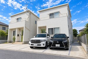 A two-story residential building with a white exterior, a driveway, and two parked SUVs. -  Grandioso Okinawa Villa Onna 5 (Onna)