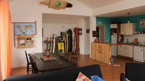 A dining area with a wooden table and chairs, a surfboard rack, and a kitchen with teal walls. - Da Mata Kite&Surf House (La Oliva)