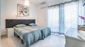 Une chambre à coucher comprenant un lit, une table de chevet, une lampe fixée au mur, une fenêtre avec un rideau et une œuvre d’art décorative accrochée au mur.