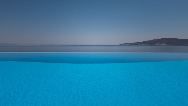 A clear view of a large, shallow pool with a gradient of blue, leading to a calm sea and a distant island. - Seafront Aegeans blue pearl over infinity pool (Keratea)