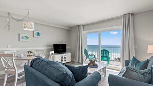 Ein modernes Wohnzimmer mit einer Couch, einem Esstisch und einem Fernseher. Ein Balkon bietet Blick auf den Ozean.