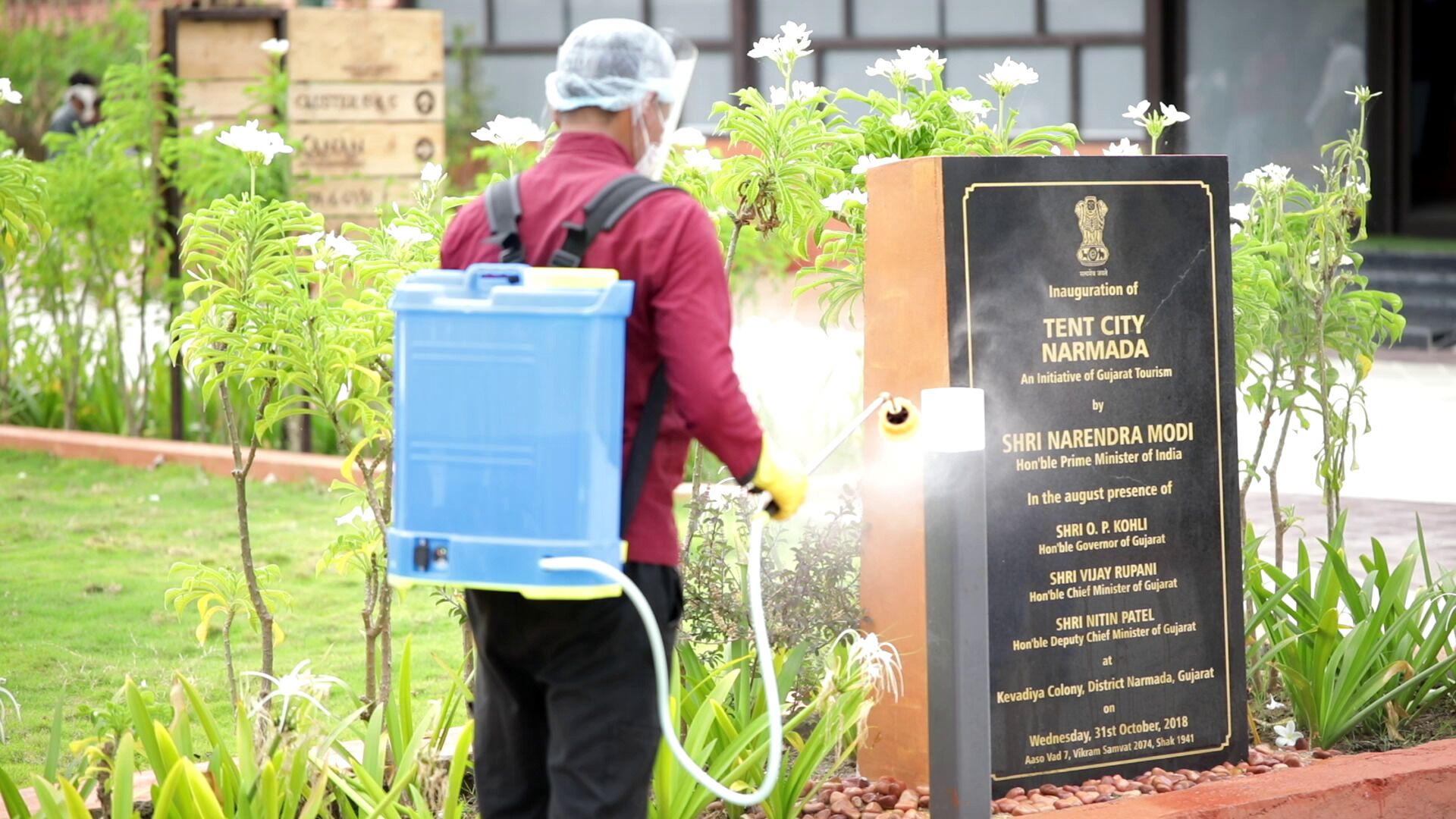 A person in a red jacket and protective gear spraying plants near a signboard.