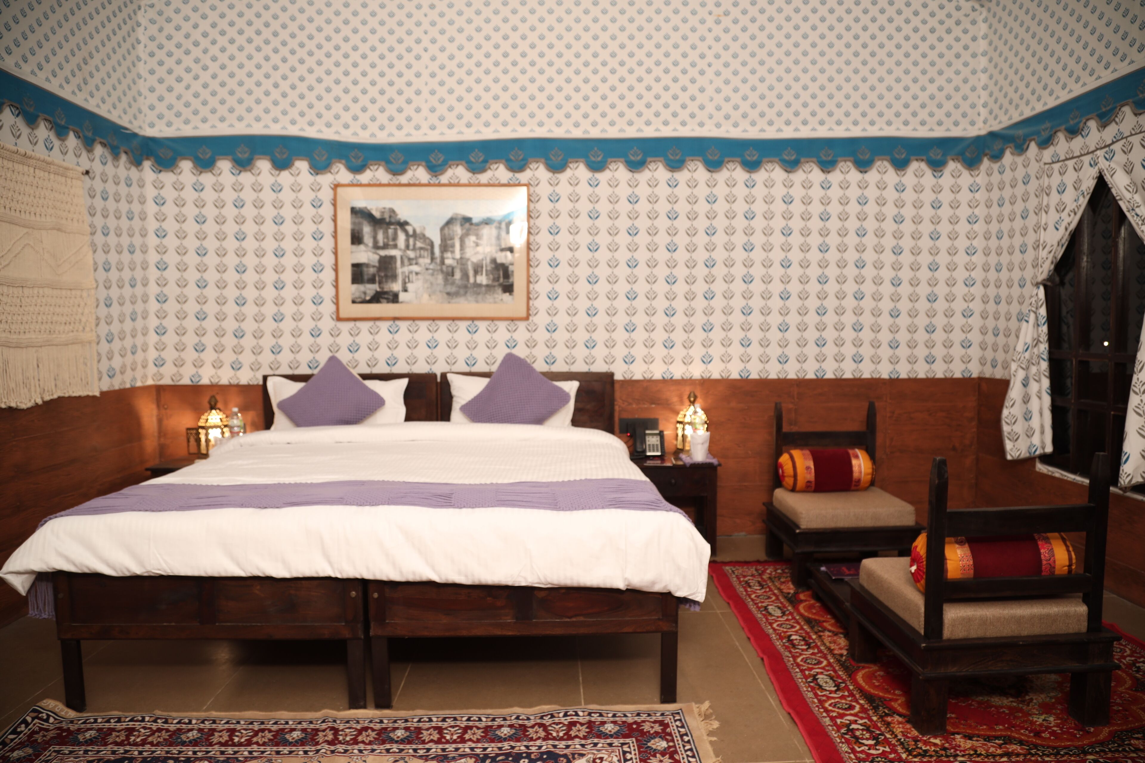 Une chambre avec un grand lit, une tête de lit en bois, un papier peint à motifs, un tableau encadré accroché au mur et un tapis aux motifs rouges et blancs.