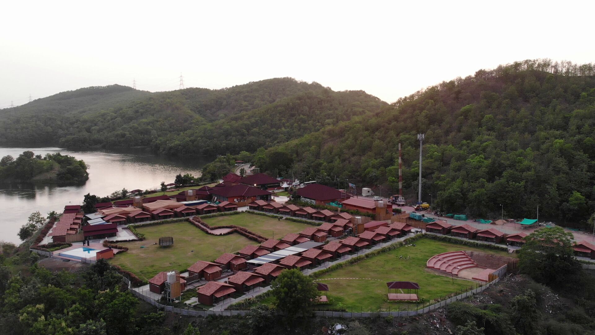 A resort with multiple red-roofed buildings arranged in a grid pattern, surrounded by lush green hills and a body of water.