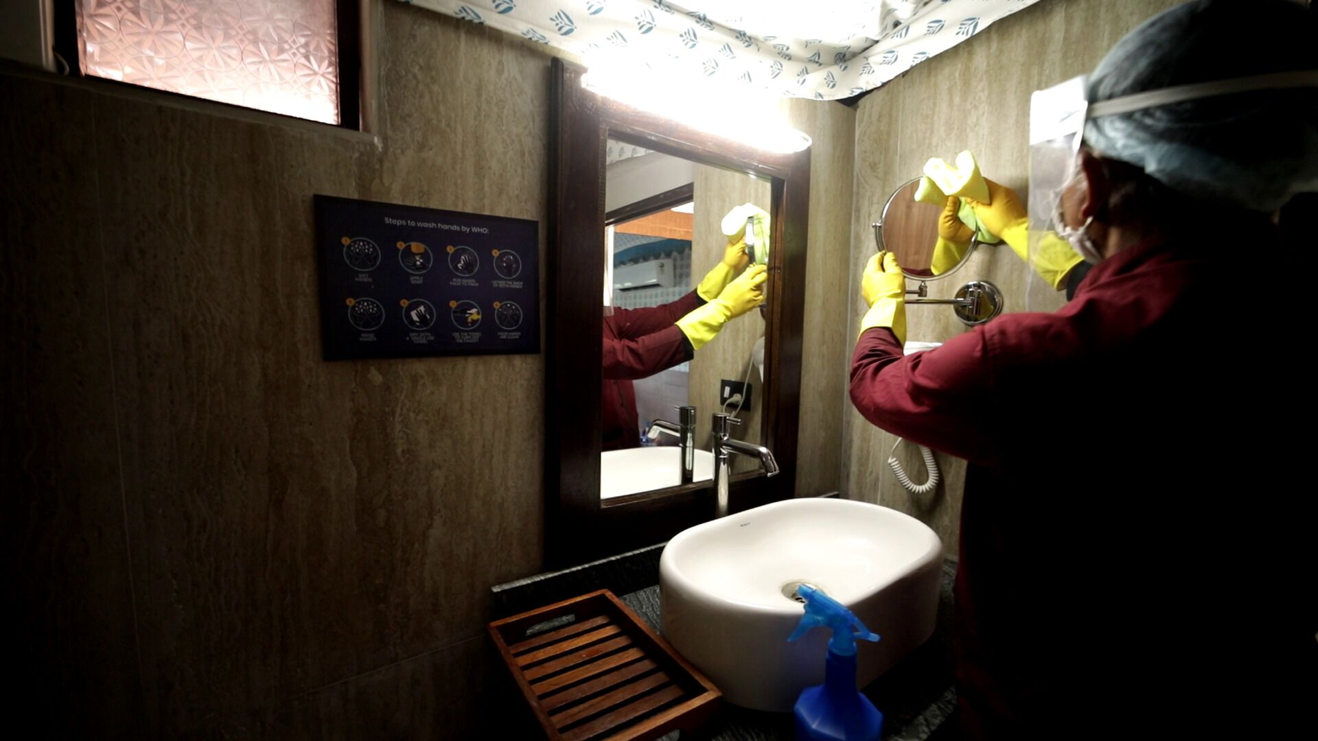 A person cleaning a sink in a bathroom with a poster on the wall and a mirror reflecting the scene.