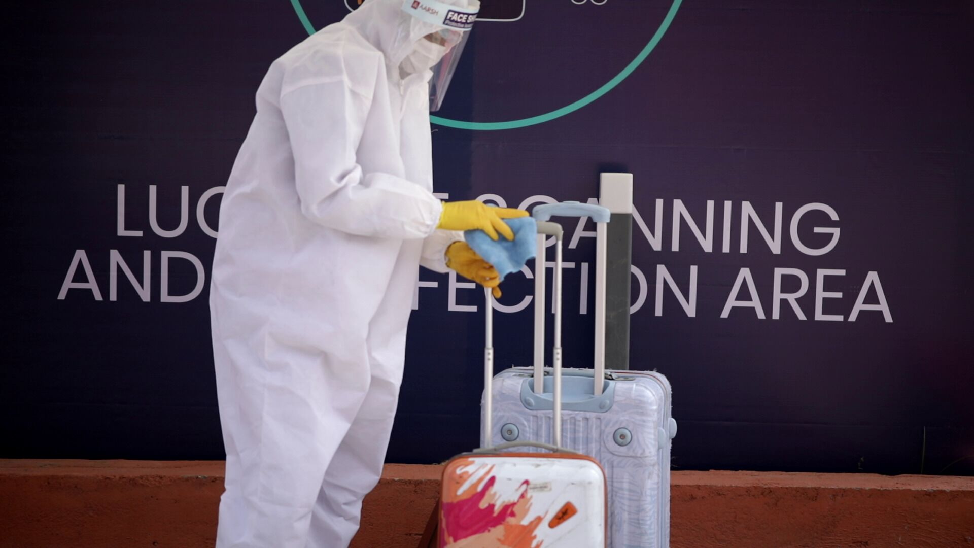 A person in a white suit and yellow gloves cleaning a suitcase in front of a sign that reads "LUCAN SCANNING AND DISINFECTION AREA".