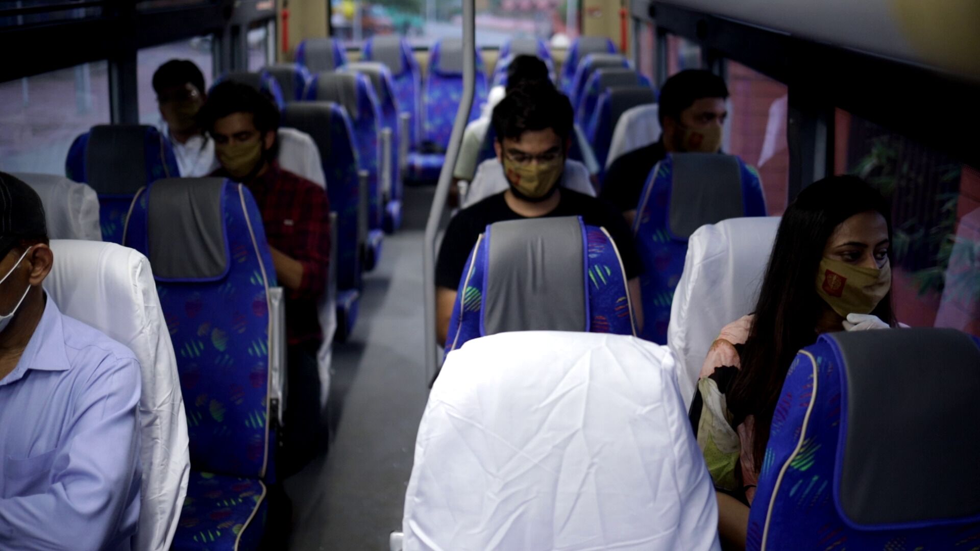 Interior de autobús con hileras de asientos, algunos cubiertos con telas blancas, y pasajeros con mascarillas.