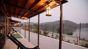A covered outdoor seating area with lounge chairs and hanging lanterns, overlooking a body of water and hills. - Statue Of Unity Tent City (Garudeshwar)