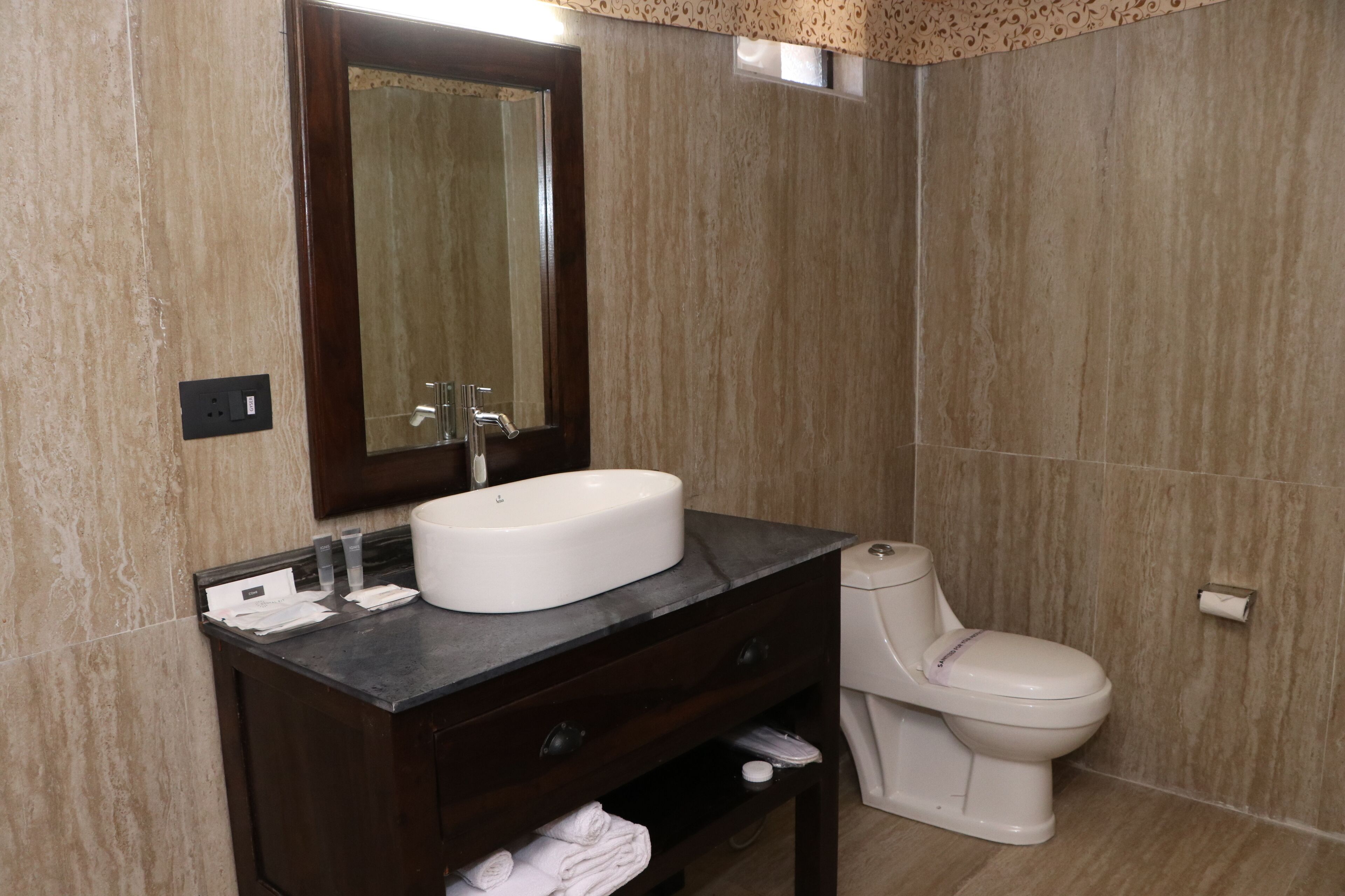 A bathroom with a marble wall, a white sink, a dark wooden vanity, a mirror, a toilet, and a towel rack.