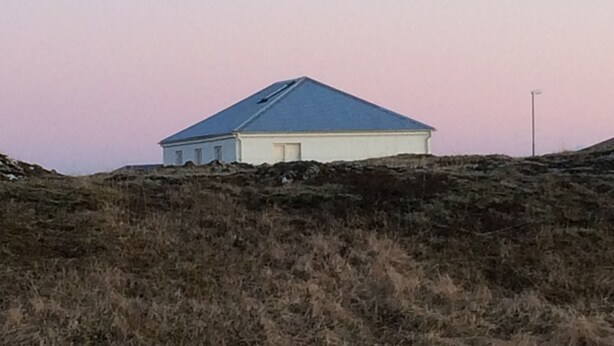 A single-story building with a sloped roof, situated on a grassy hill. - Skipholt (Vogar)