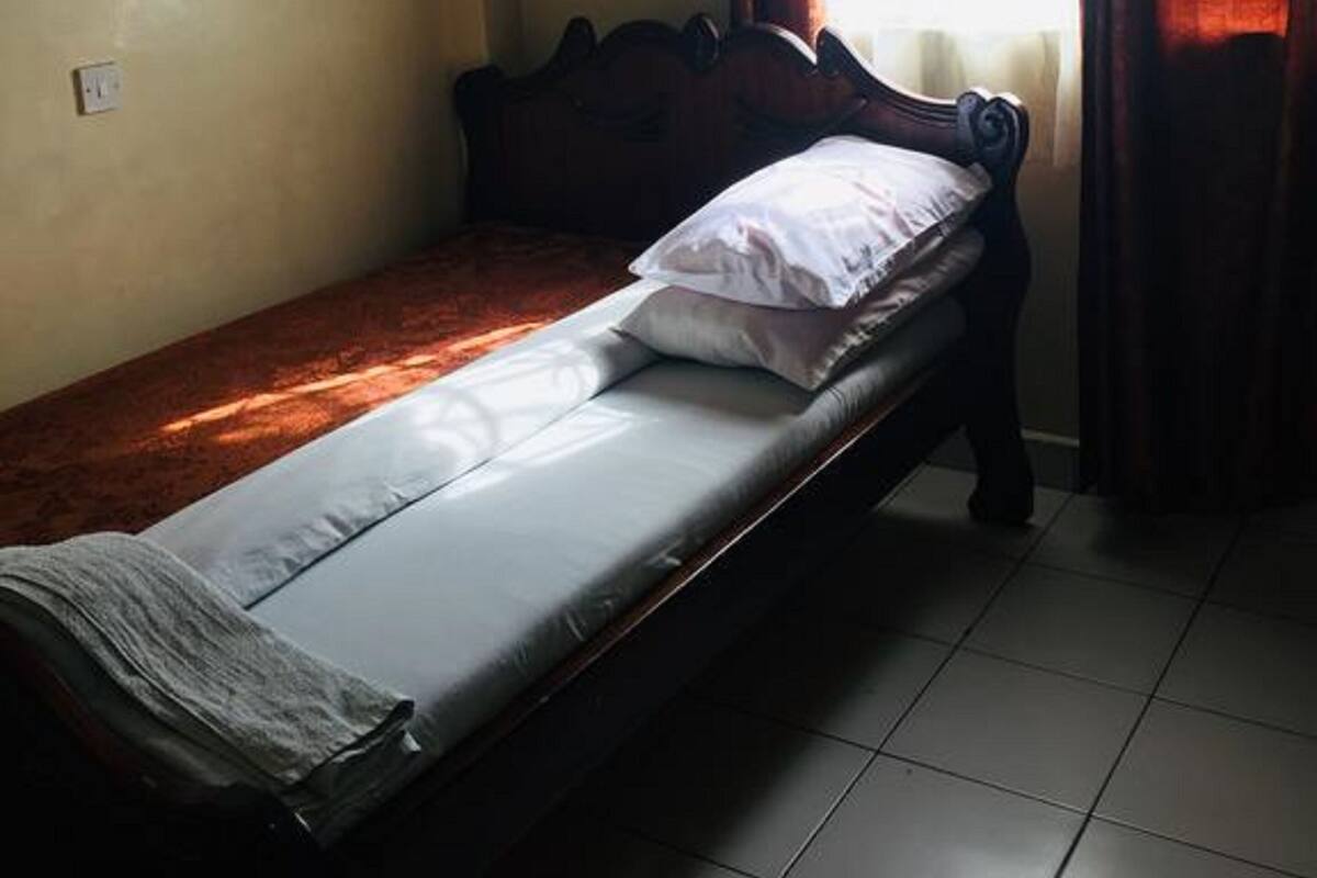 Un lit simple avec une tête de lit en bois, un oreiller blanc et une couverture. Il y a une fenêtre avec des rideaux, et une prise de courant est visible sur le mur.