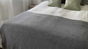 Una cama bien hecha con una manta gris y almohadas, una lámpara de noche y una ventana con cortinas translúcidas.