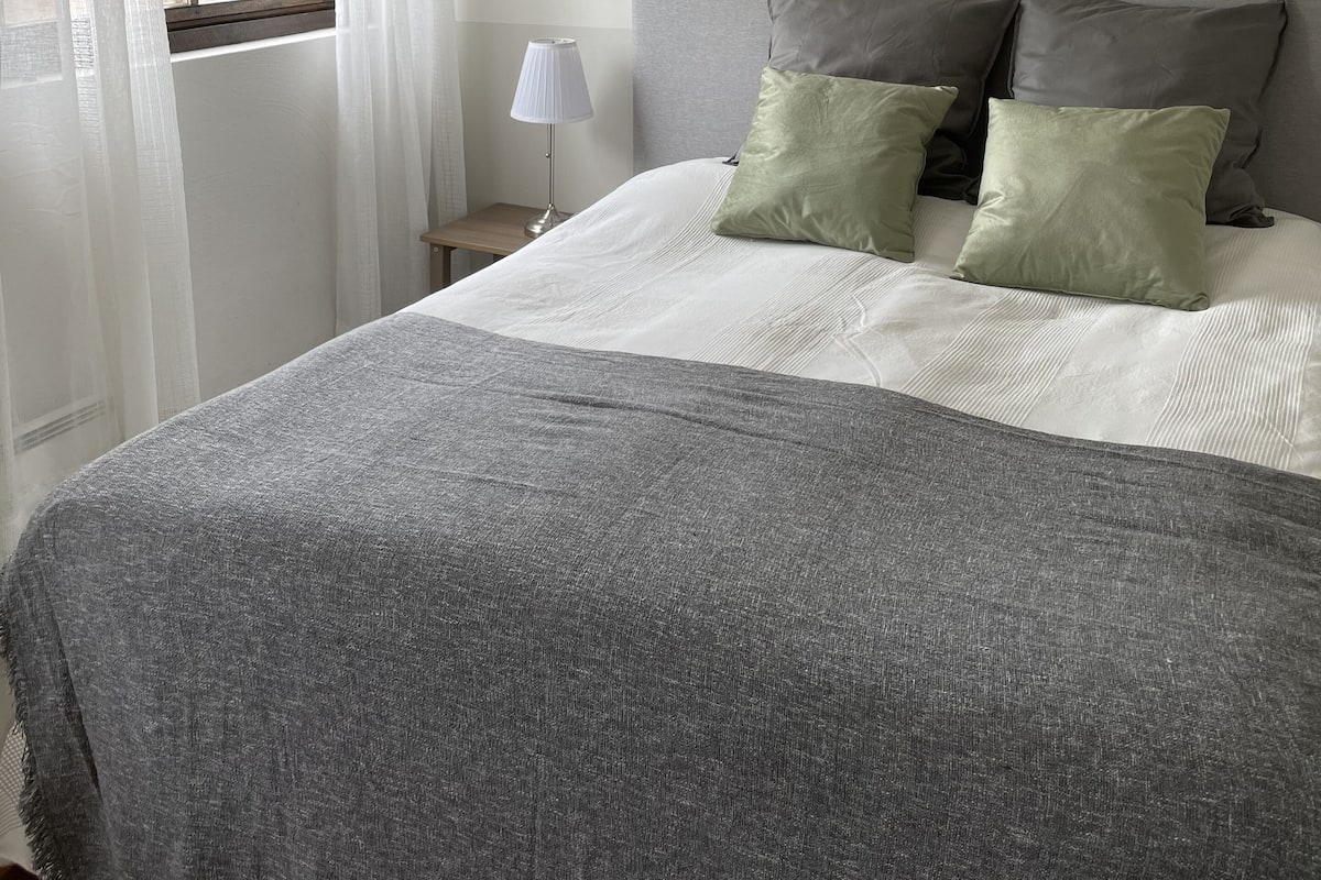 Una cama bien hecha con una manta gris y almohadas, una lámpara de noche y una ventana con cortinas translúcidas.