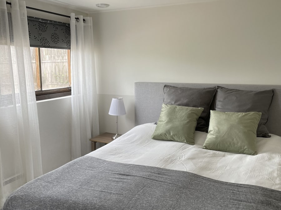 Una cama bien hecha con una manta gris y almohadas, una lámpara de noche y una ventana con cortinas translúcidas.