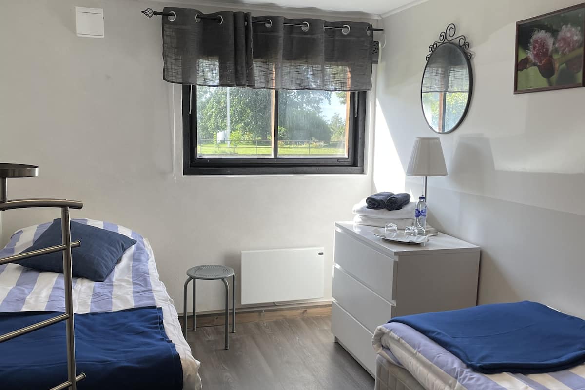 Un dormitorio con dos camas, un aparador, un radiador, una ventana con cortinas, un espejo y un taburete pequeño.
