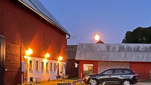 Un edificio con fachada roja y un coche negro estacionado adelante. Hay áreas de estar al aire libre con mesas y sillas.