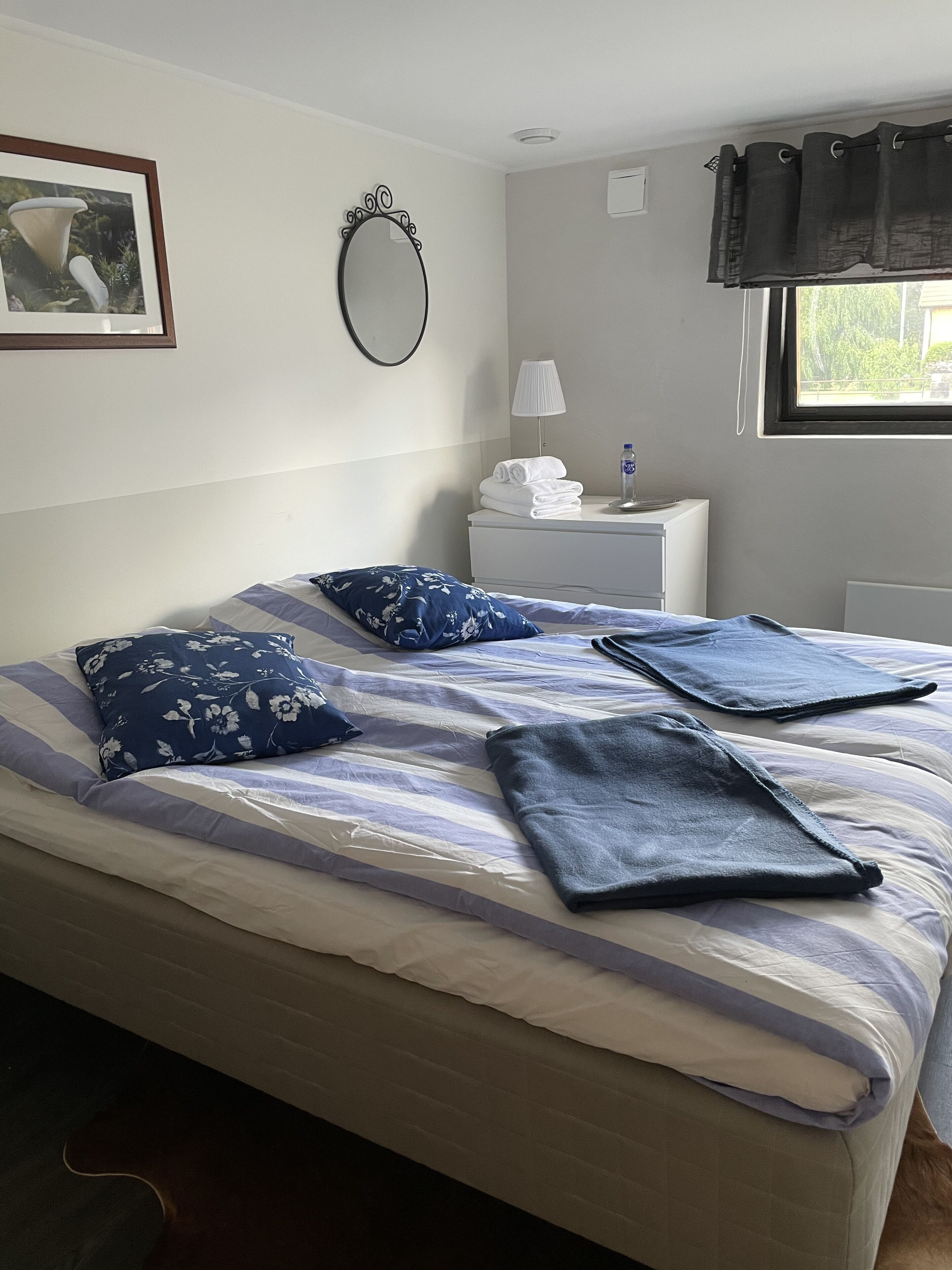 A neatly made bed with a striped bedspread and two folded blankets, a bedside table with a lamp and a bottle, a framed picture on the wall, and a window with curtains.