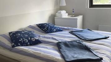 A neatly made bed with a striped bedspread and two folded blankets, a bedside table with a lamp and a bottle, a framed picture on the wall, and a window with curtains.