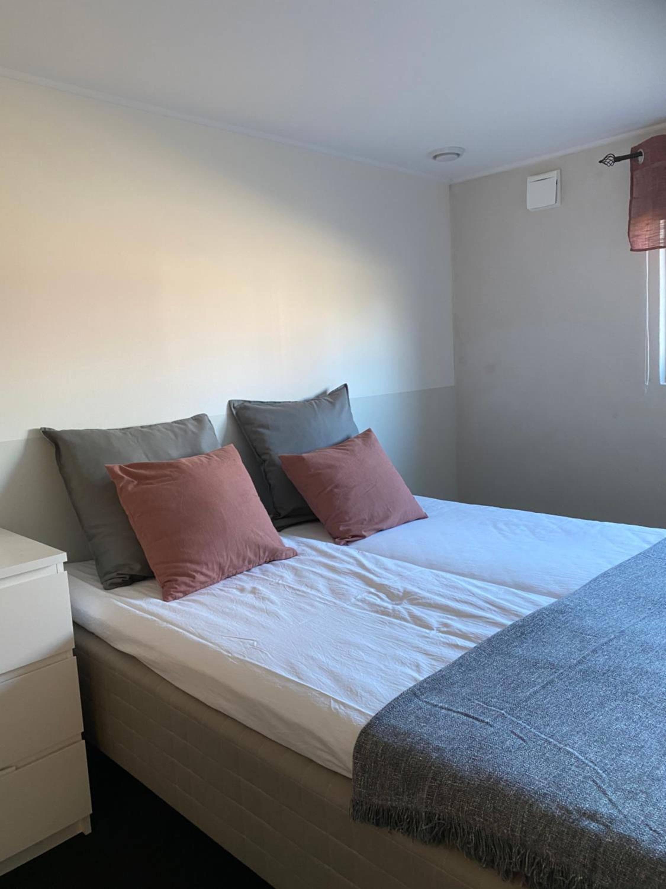A single bed with a white mattress and two pillows, a grey blanket, and a white nightstand with drawers.
