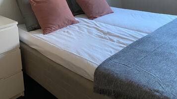 A single bed with a white mattress and two pillows, a grey blanket, and a white nightstand with drawers.