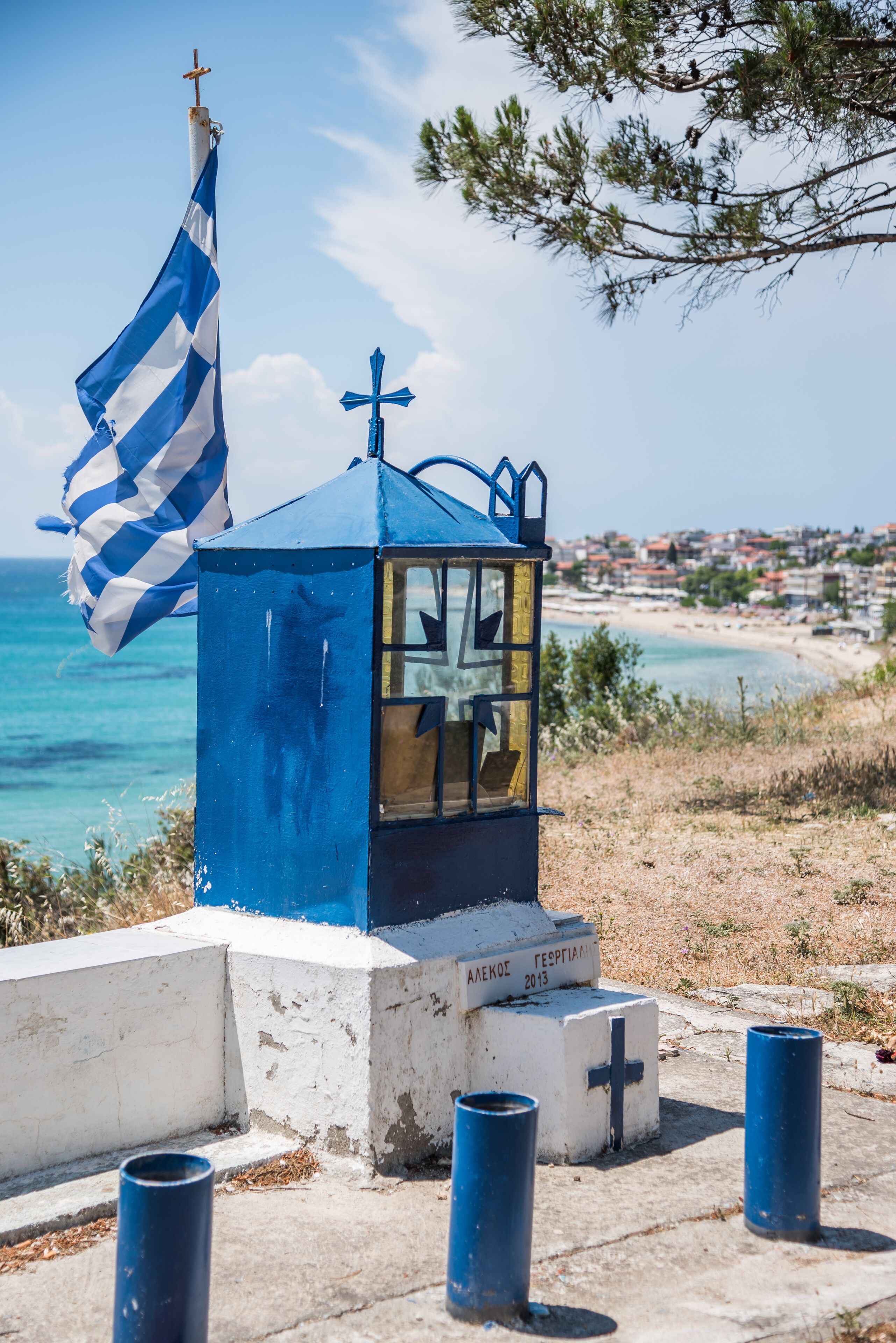 A small blue and white structure with a flag, a cross, and a plaque reading "ΑΛΕΞΟΙ 2013" near a beach.