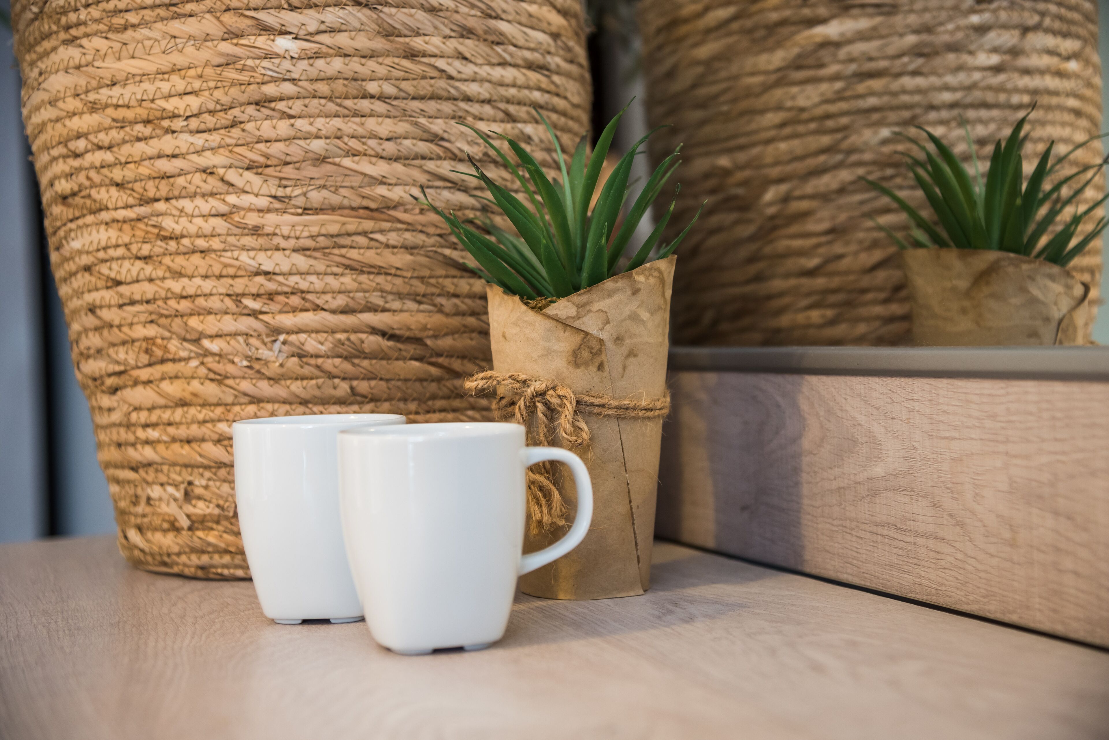A rustic setting with a woven basket, a small potted plant, and two white mugs on a wooden surface.