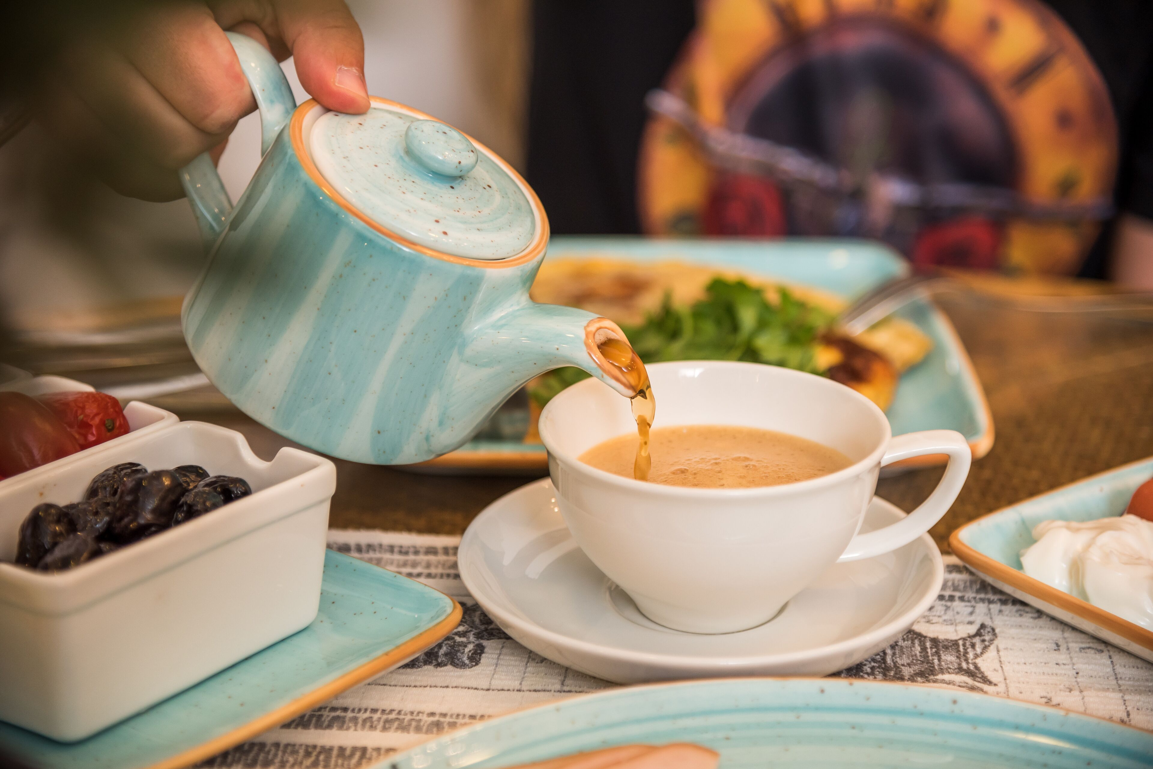 Una mano sirve café en una taza sobre una mesa con varios platos y un periódico.