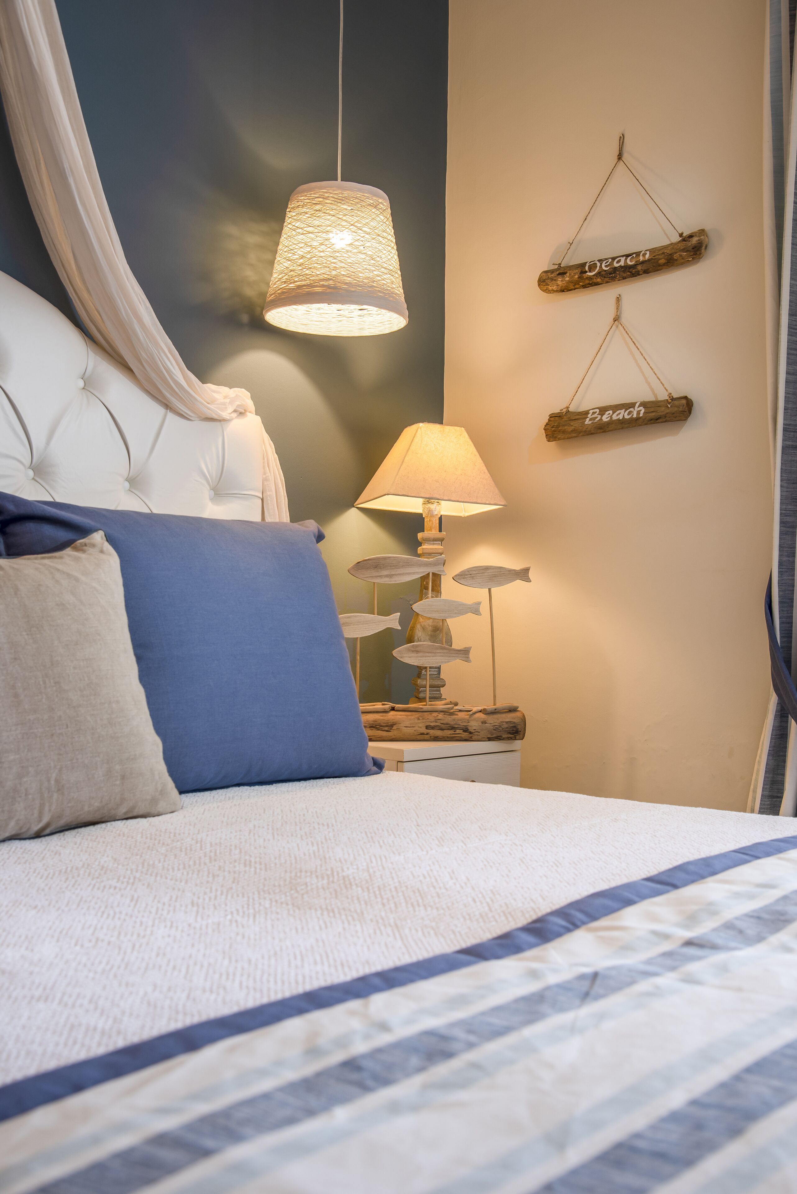 Een slaapkamer met een gestoffeerd hoofdeinde, een bed met gestreepte beddengoed, een hanglamp, een nachtkastje met een lamp en decoratieve objecten, en aan de muur bevestigde bordjes met de tekst 'Strand'.