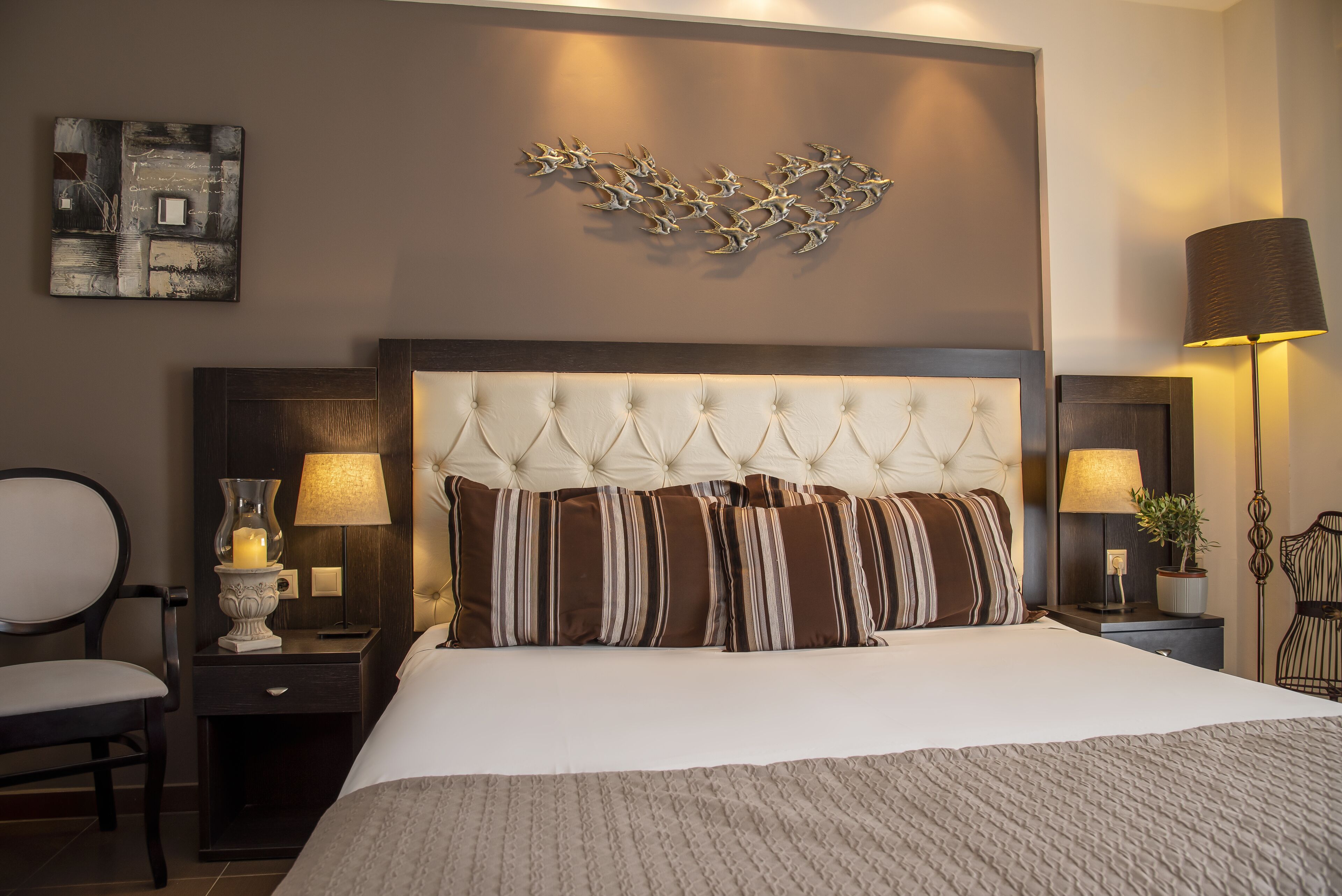 Habitación de hotel con una cama grande, cabecero acolchado, mesita de noche con lámpara, una silla y una obra de arte en la pared.