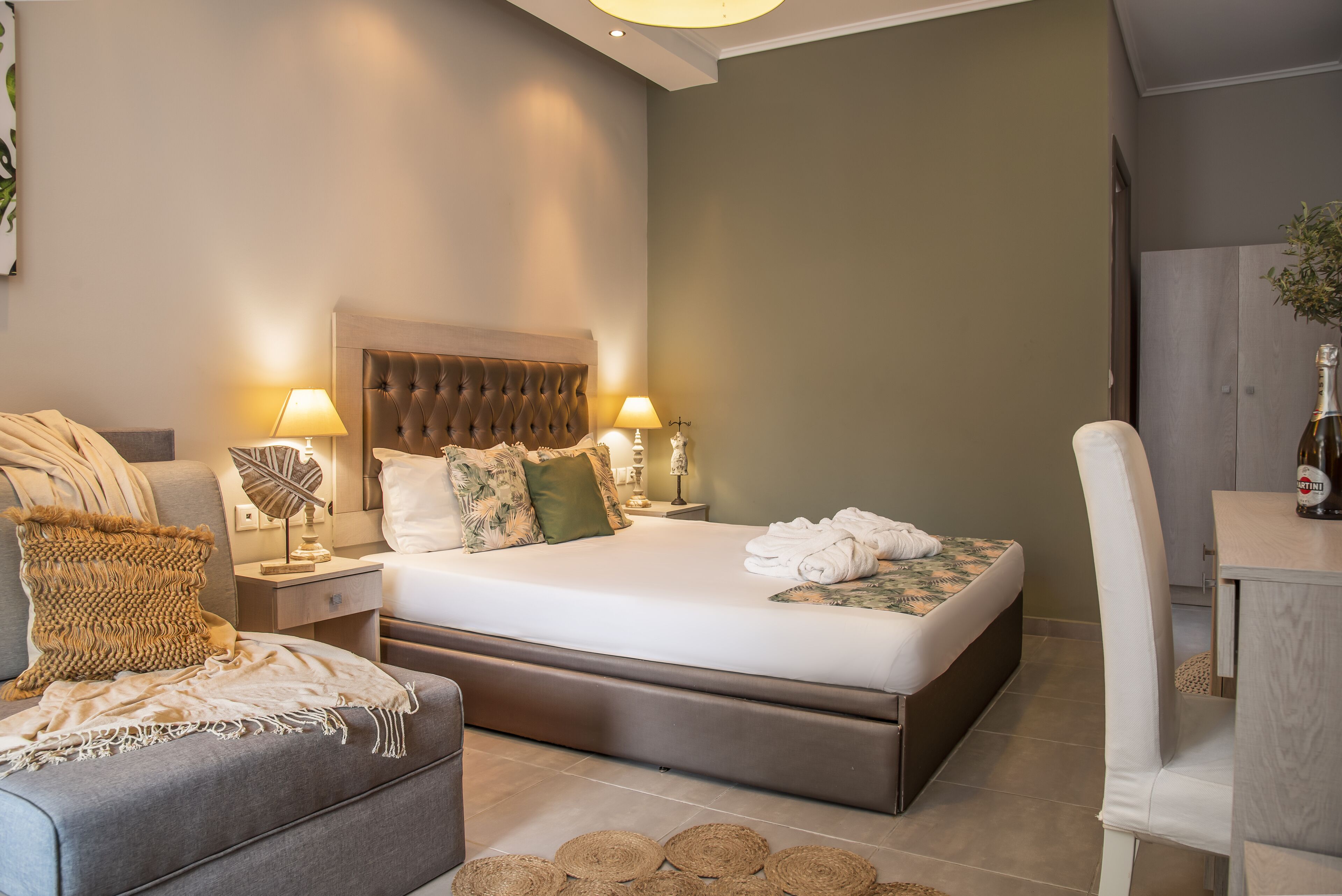 A modern bedroom with a large bed, two bedside tables with lamps, a grey sofa, and a wooden chair.
