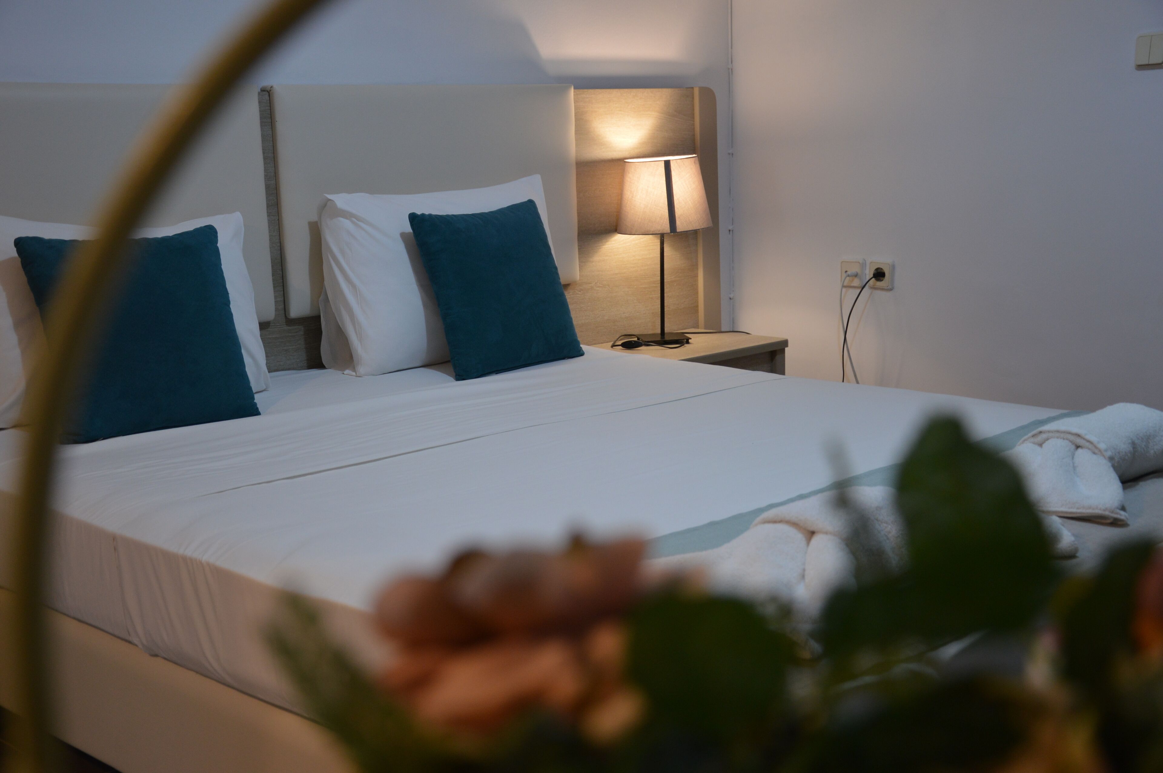 Una cama bien hecha con sábanas blancas y dos almohadas turquesa, una mesita de noche con lámpara y un enchufe en la pared.