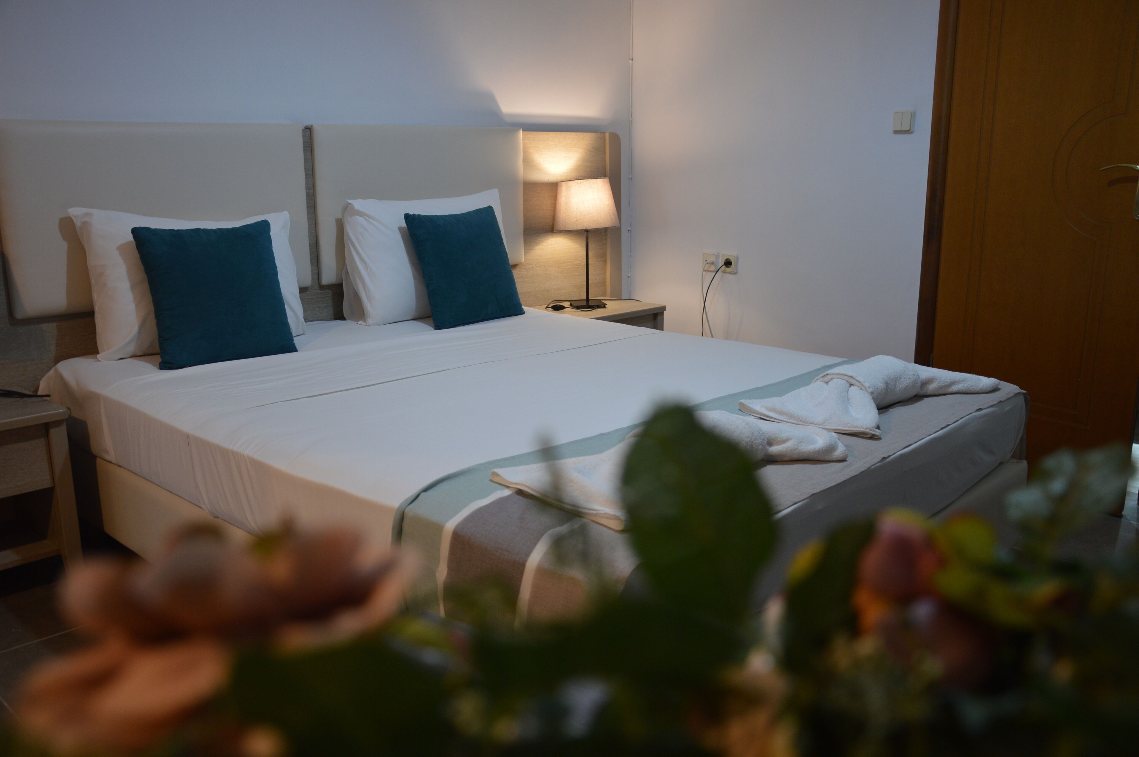 Una cama bien hecha con almohadas blancas y azul verdosas, una lámpara de noche y una mesita con una lámpara.