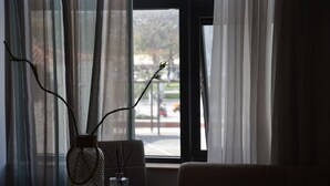 A room with a table and chair, a vase with branches, and a view of a street through a window with sheer curtains. - Aqua Mare Sea Side (Nea Propontida)