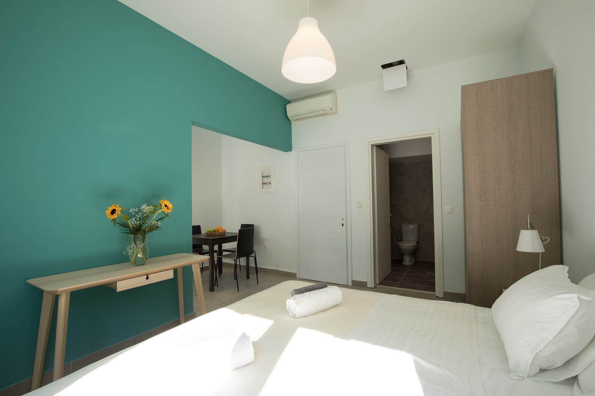 A modern hotel room with a bed, a desk, a chair, a dining table, and a bathroom visible through an open door.