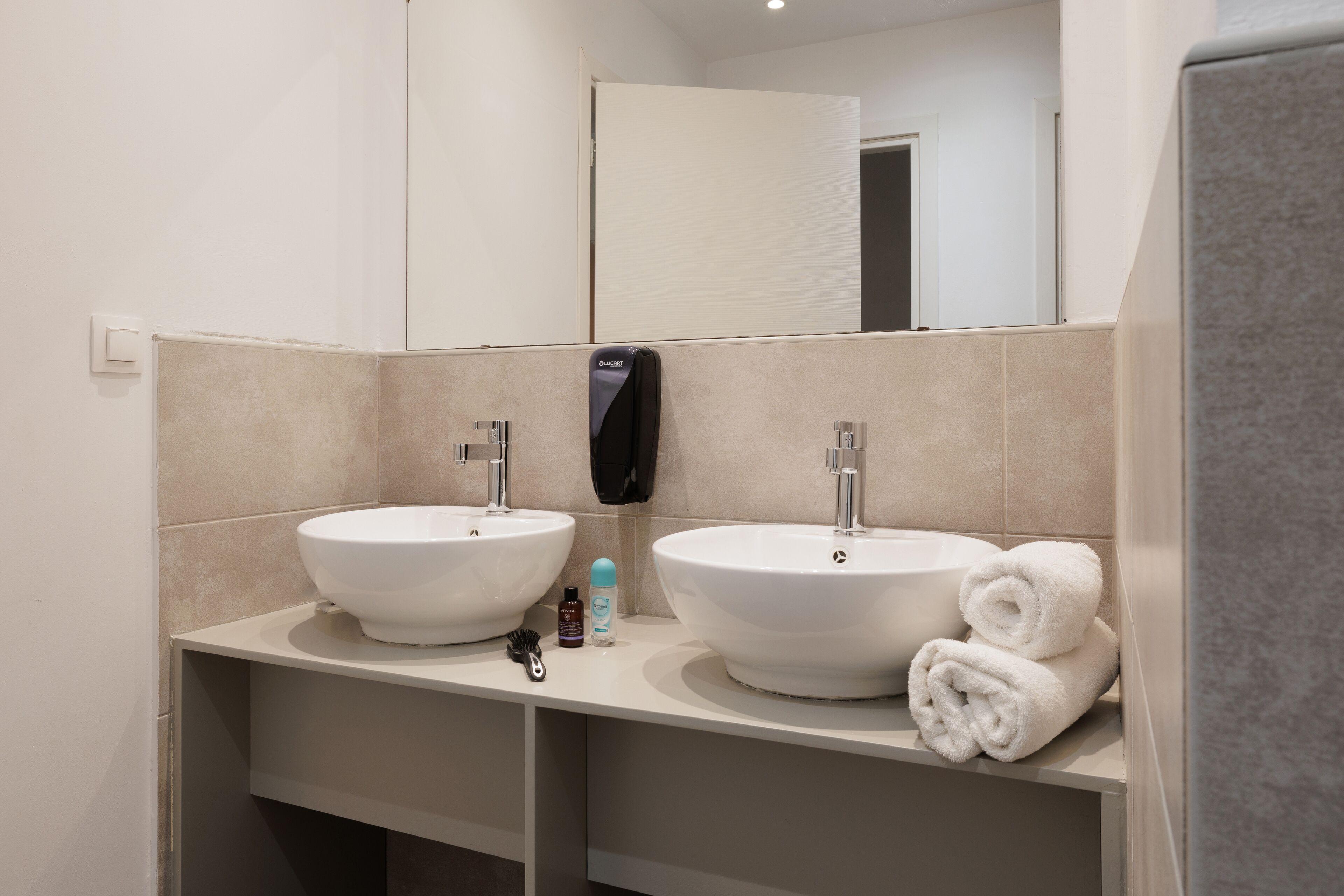 A bathroom with two sinks, a mirror, a towel rack, and a soap dispenser.