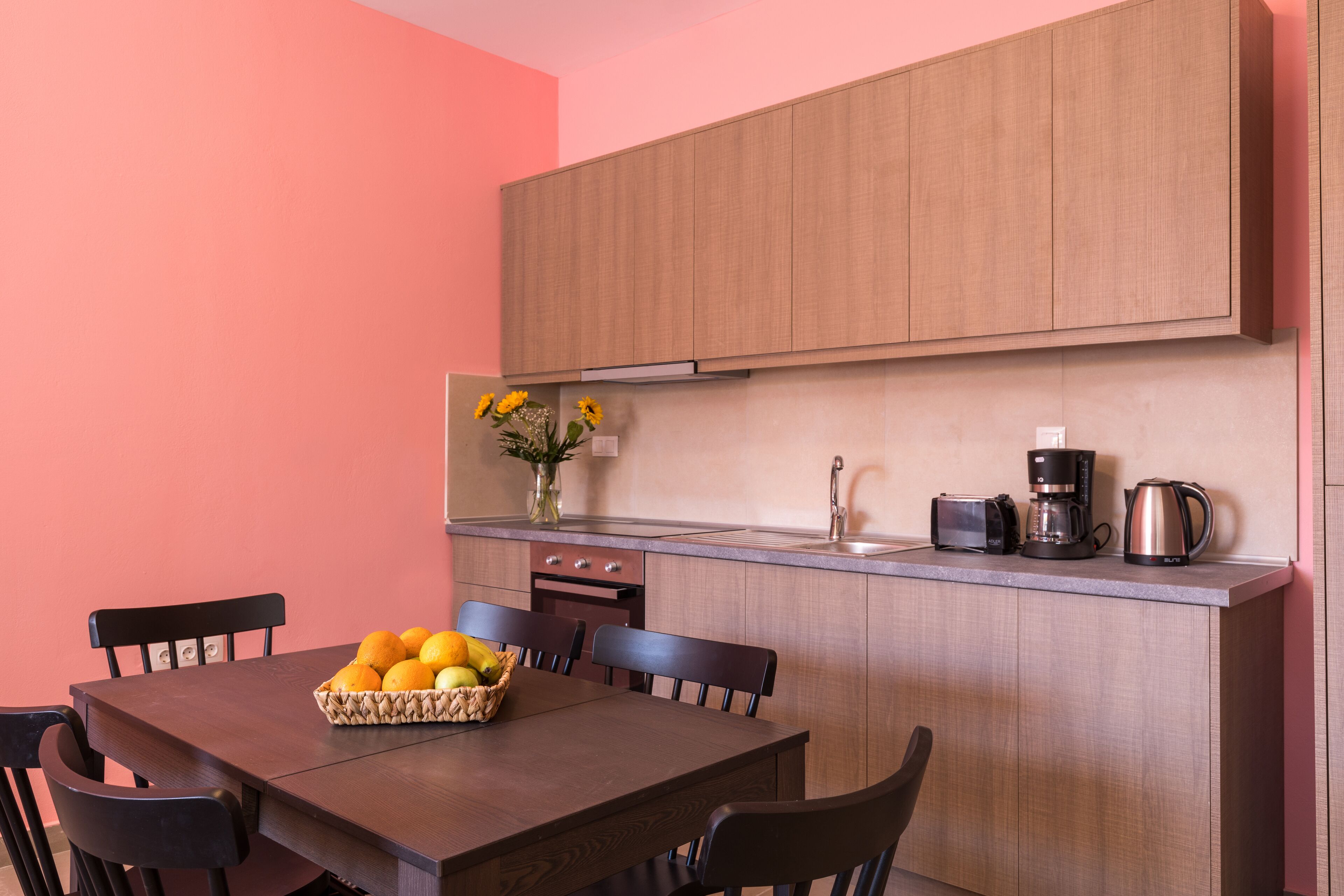 A modern kitchen with wooden cabinets, a dining table with chairs, and a fruit basket.