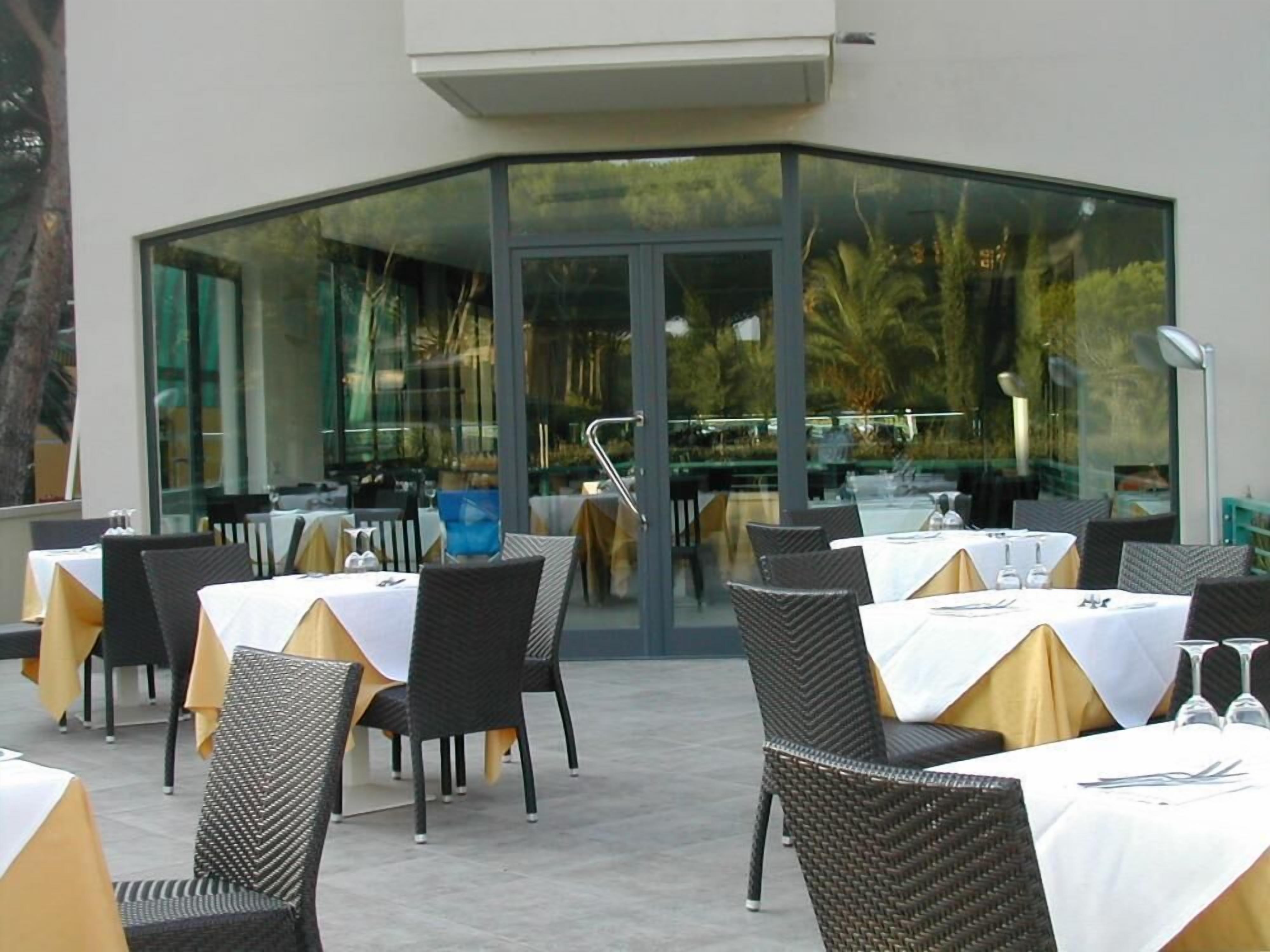 An outdoor dining area with tables and chairs, a glass enclosure, and a view of the surrounding greenery.