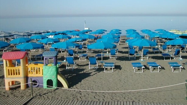 A beach with rows of blue umbrellas and lounge chairs, a wooden boardwalk, and colorful play structures. - Hotel Flora (Bibbona)