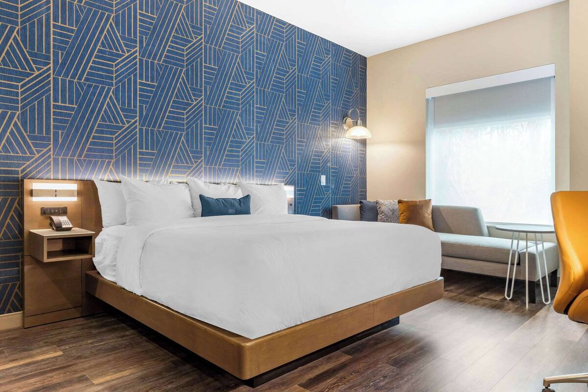 A modern hotel room with a large bed, a nightstand with a lamp, a seating area with a sofa and armchairs, and a large window with a view.