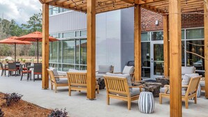 Un patio al aire libre con mobiliario de madera y sombrillas, con vistas a un edificio moderno con grandes ventanales.