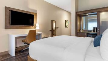 A hotel room with a bed, a desk with a chair, a television mounted on the wall, a mirror, and a coffee machine.