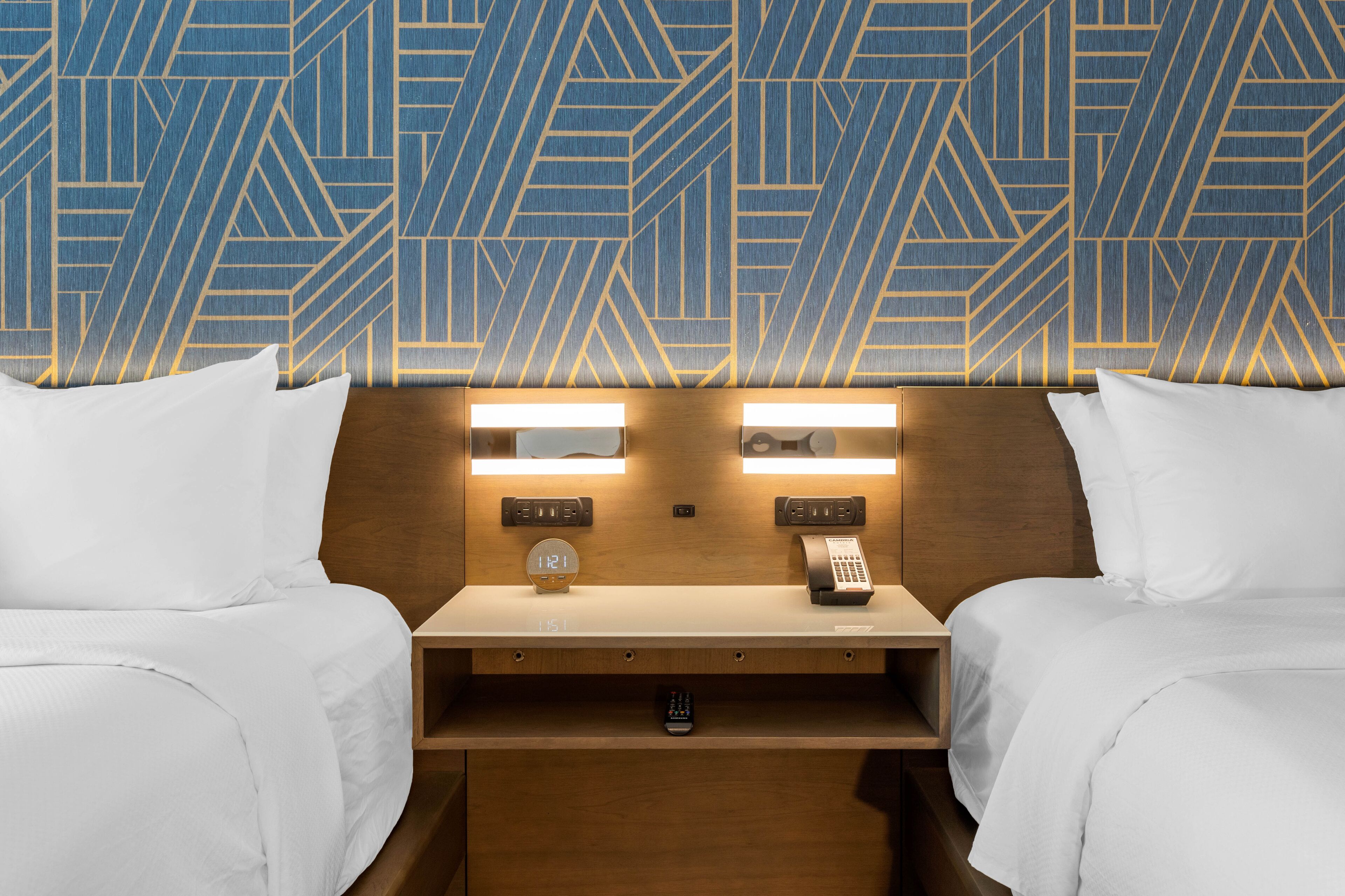 Habitación de hotel con dos camas, una mesita de noche con un reloj y un teléfono, y una pared con un diseño geométrico.