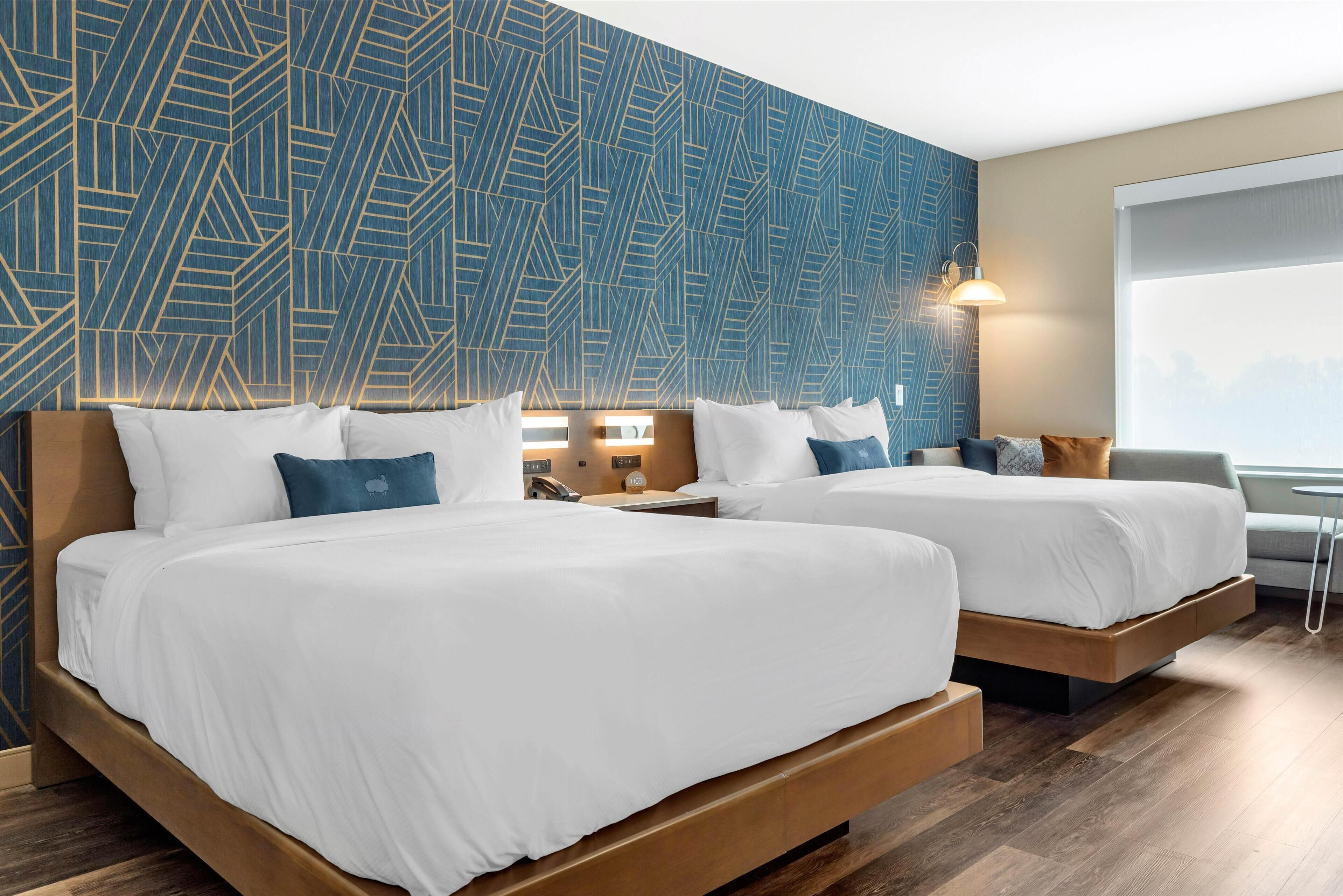 Quarto de hotel com duas camas, parede estampada, mesa de cabeceira, abajur e janela.