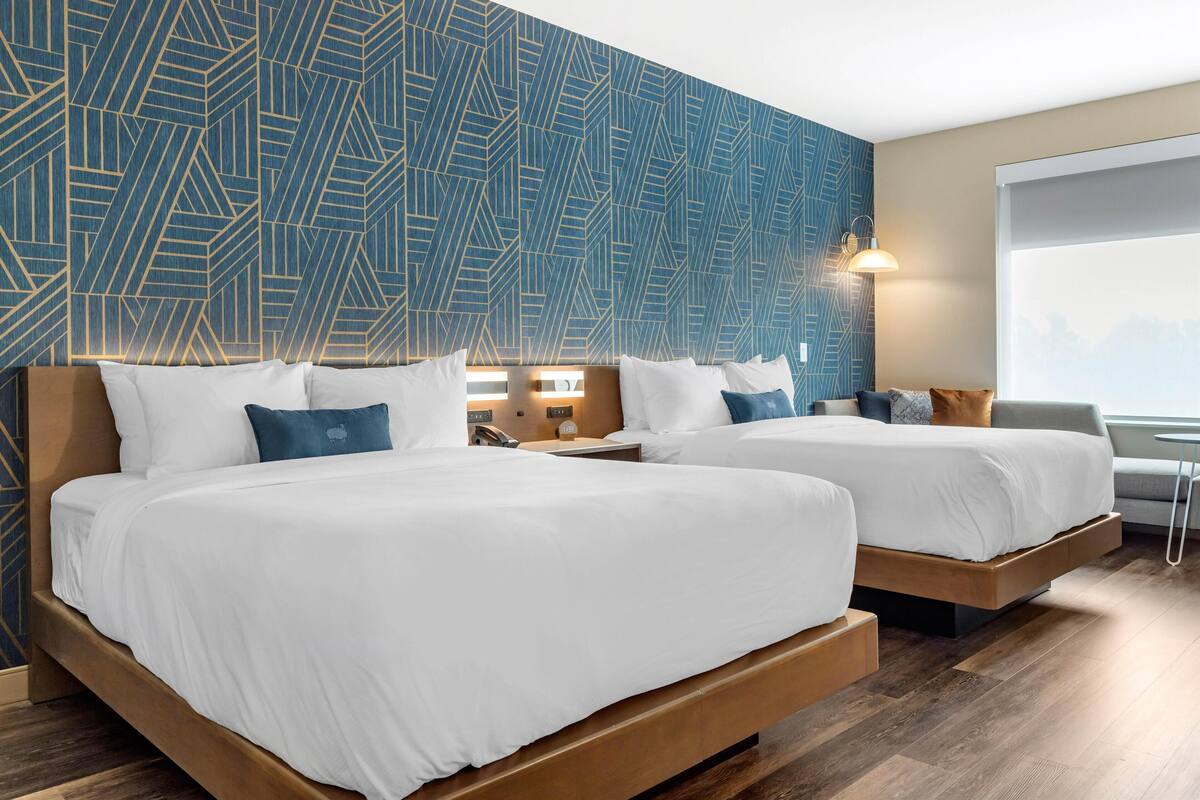 A hotel room with two beds, a patterned wall, a nightstand, a lamp, and a window.