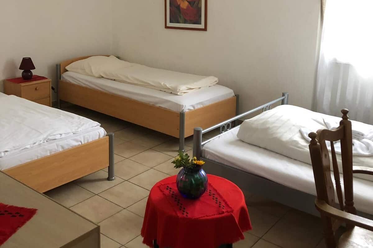 A small, clean room with two single beds, a small table with a vase, and a chair.